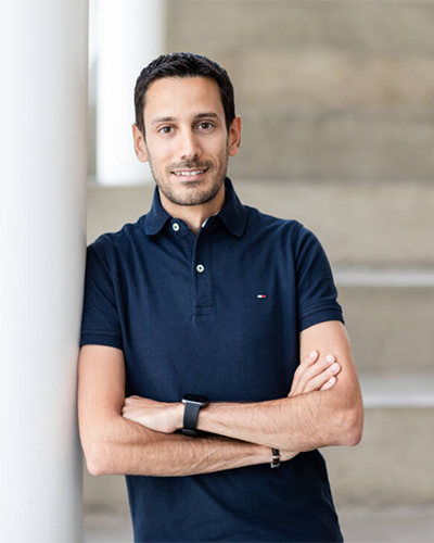 Portrait d'un homme souriant en polo bleu marine, adossé à une colonne, mettant en valeur une atmosphère professionnelle et conviviale.