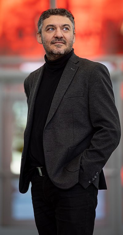 A confident man, dressed in a dark suit, stands in front of a bright and modern background, evoking a dynamic academic environment.