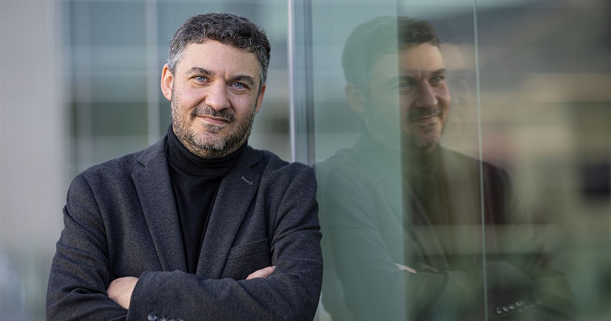 Un homme souriant, en manteau sombre, pose avec confiance devant une surface vitrée réfléchissante. L'ambiance est professionnelle et claire.