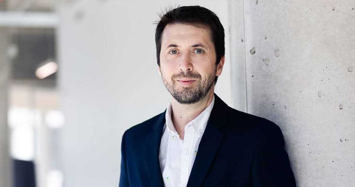 A smiling man, dressed in a blazer, stands against a minimalist wall, expressing confidence and professionalism.