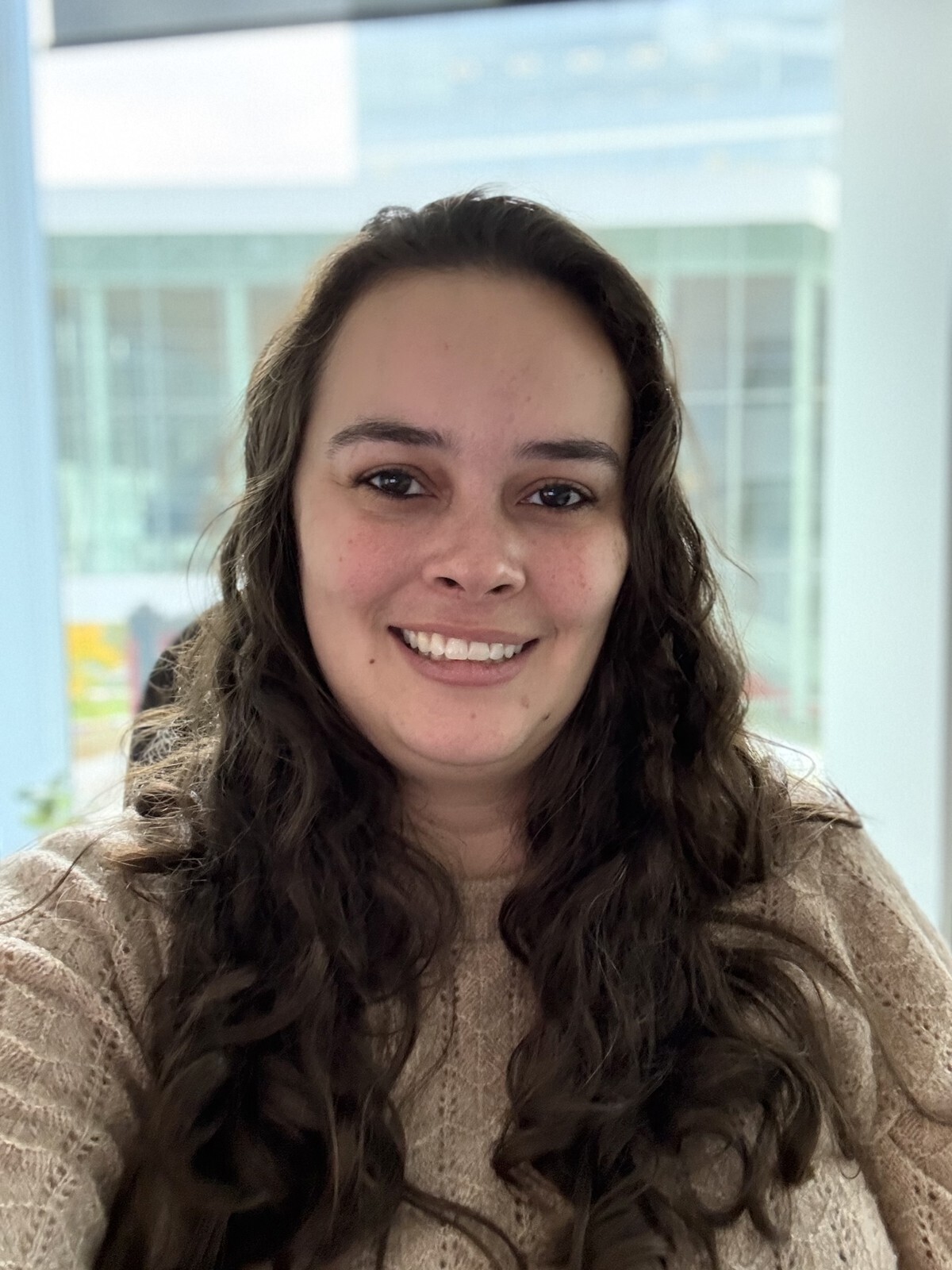Étudiante souriante, cheveux bouclés et pull beige, au sein d'un environnement lumineux et moderne, symbole de l'innovation technologique.
