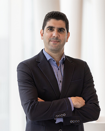 Un professionnel souriant, vêtu d'un costume sombre, se tient debout avec les bras croisés, dans un cadre moderne et lumineux.