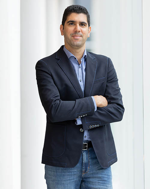 Un professionnel souriant, vêtu d'un blazer noir et d'une chemise bleu, se tient debout devant une grande fenêtre.