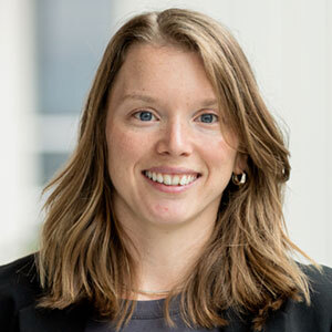 Portrait d'une professionnelle souriante, aux cheveux longs et châtains, vêtue de manière formelle, reflétant dynamisme et engagement.
