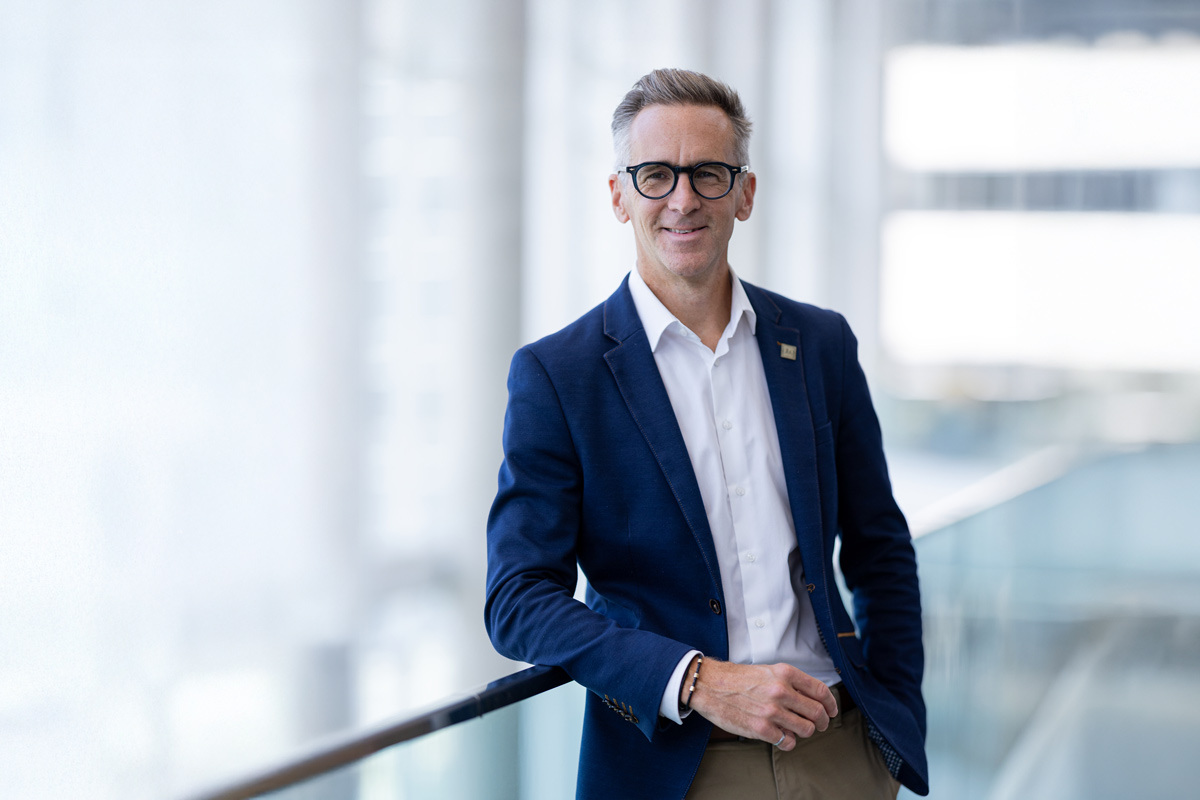 Un professionnel souriant, vêtu d'un blazer, pose avec confiance dans un environnement lumineux et moderne.