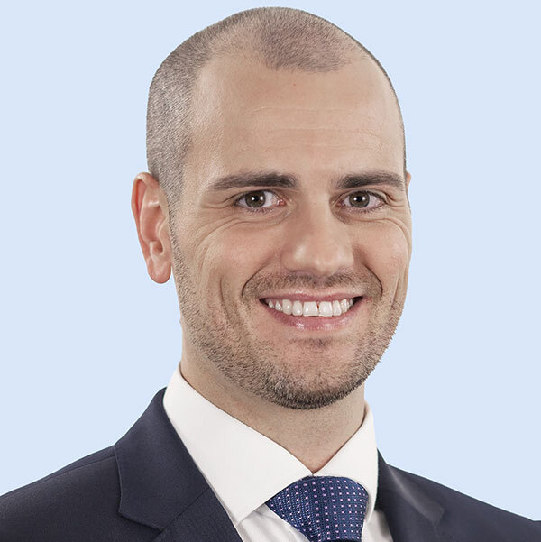A professional individual wearing a suit, smiling confidently against a light blue background.