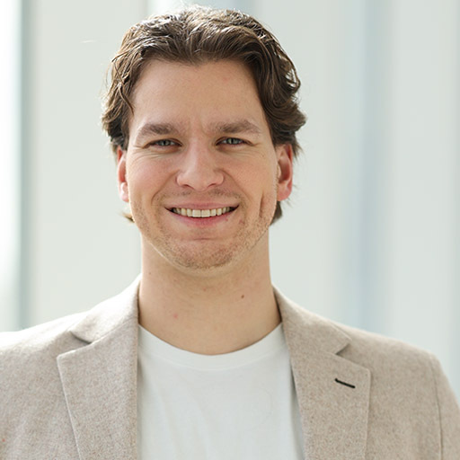 Un jeune professionnel souriant, vêtu d'un blazer clair sur un t-shirt, dégage confiance et accessibilité dans un environnement moderne.