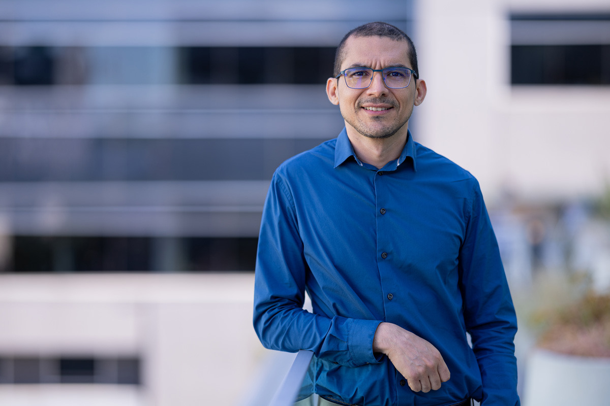 Un homme souriant porte une chemise bleu et des lunettes, en arrière-plan des bâtiments modernes d'une université.