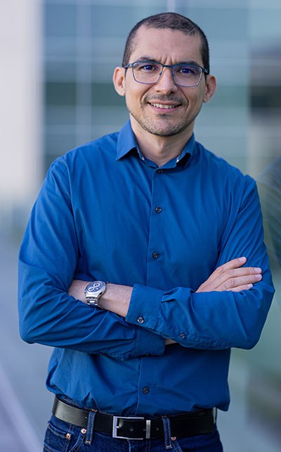 Un homme souriant en chemise bleue, les bras croisés, se tient devant un fond moderne, dégageant confiance et professionnalisme.