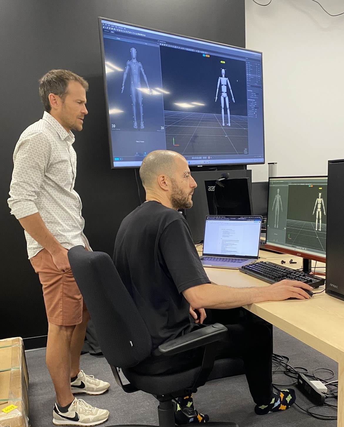 Deux personnes travaillent sur des projets de modélisation 3D, avec des écrans affichant des figures humanoïdes et des lignes de code.