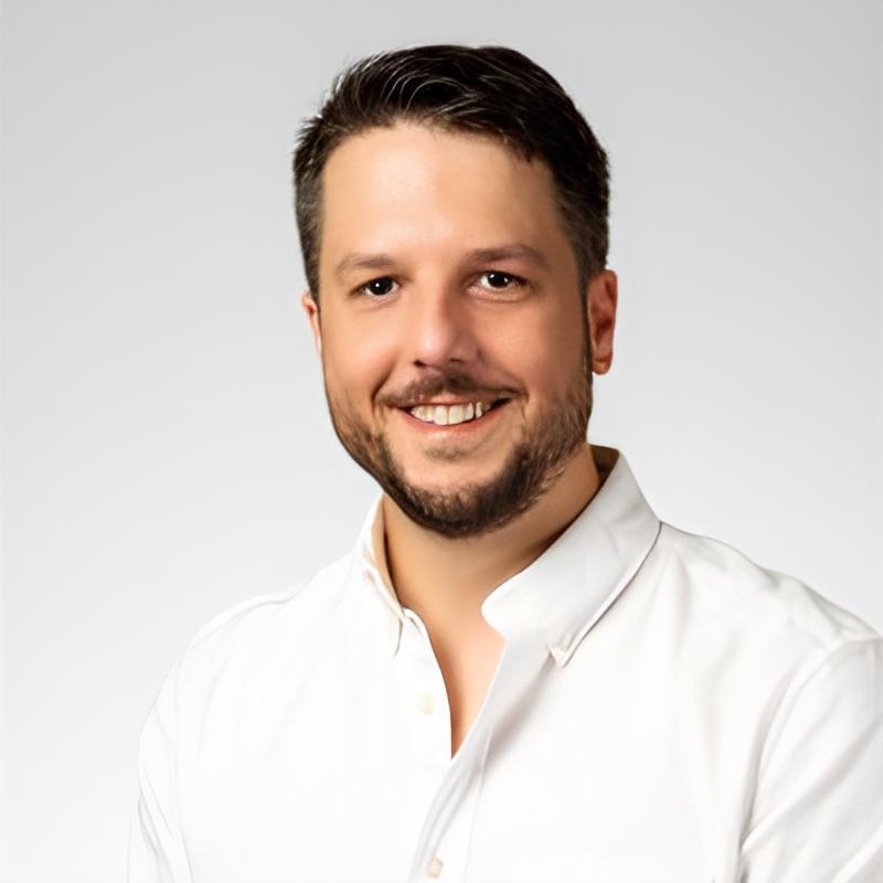 Un homme souriant, avec une barbe légère, portant une chemise blanche. Fond neutre met en valeur sa posture confiante et amicale.