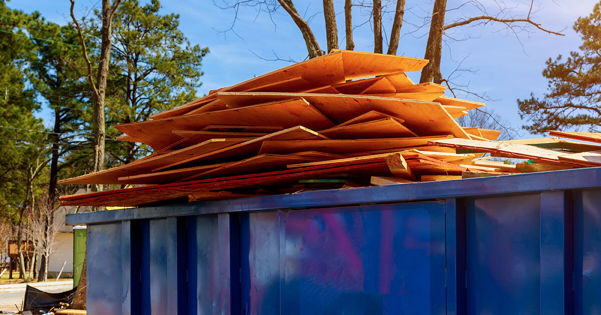 A colorful dumpster filled with stacked wooden sheets, surrounded by trees under a clear blue sky.