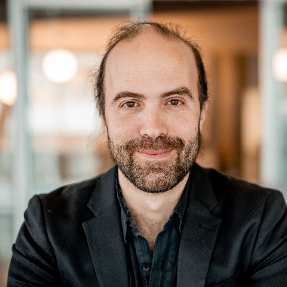 Professeur souriant, vêtu d'un blazer noir, dans un cadre moderne. Son regard amical reflète une passion pour l'enseignement.