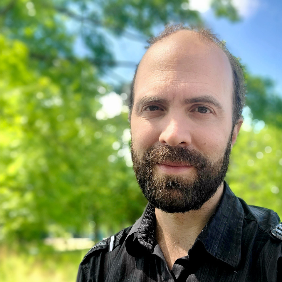 Un homme souriant avec une barbe, portant une chemise noire, se tient devant un fond verdoyant et ensoleillé.