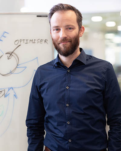 Un enseignant en technologie, souriant, devant un tableau où figurent des concepts liés à l'optimisation et à l'innovation.