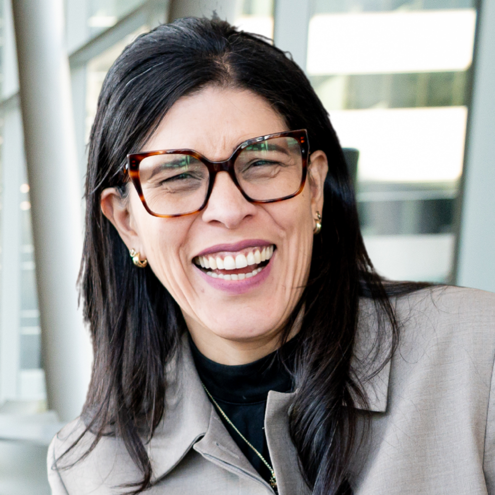 A smiling woman with long dark hair and stylish glasses, dressed in a professional outfit, exudes warmth and approachability.