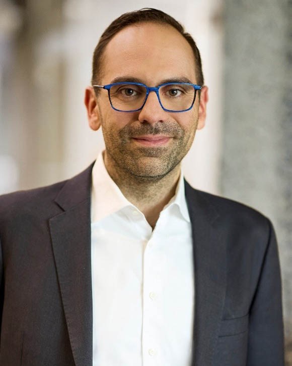 Un homme souriant portant des lunettes bleues, vêtu d'un costume sombre et d'une chemise blanche, se tient dans un intérieur moderne.