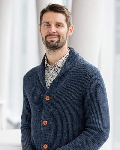 Portrait d'un homme souriant, portant un pull en laine et une chemise à motifs, dans un environnement lumineux et moderne.
