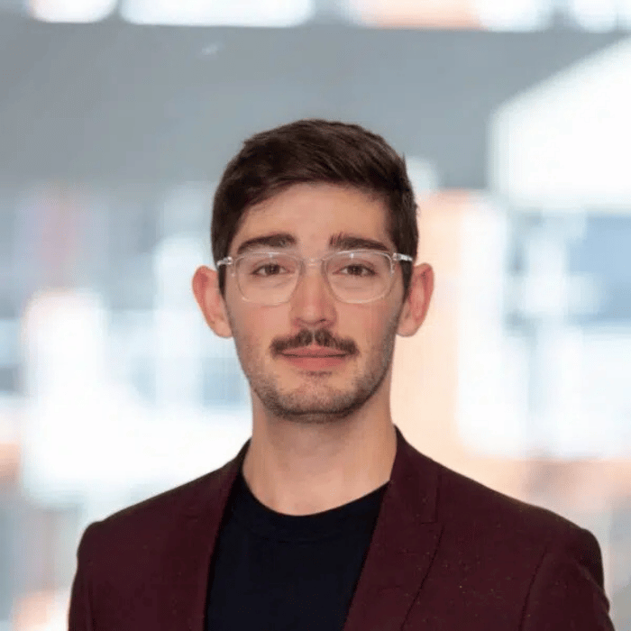 Un jeune homme avec des lunettes, un léger sourire, portant une veste foncée, se tient devant un fond lumineux.