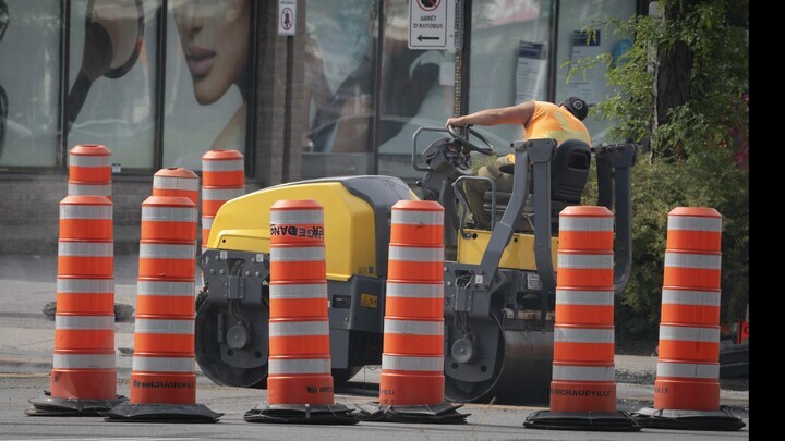 Un conducteur opère un rouleau compresseur entre des cônes de signalisation sur un chantier urbain.