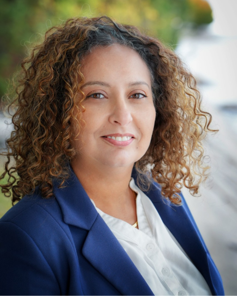 Professionnelle souriante, portant une veste bleue et une chemise blanche, se tenant devant un mur etouré de verdure.