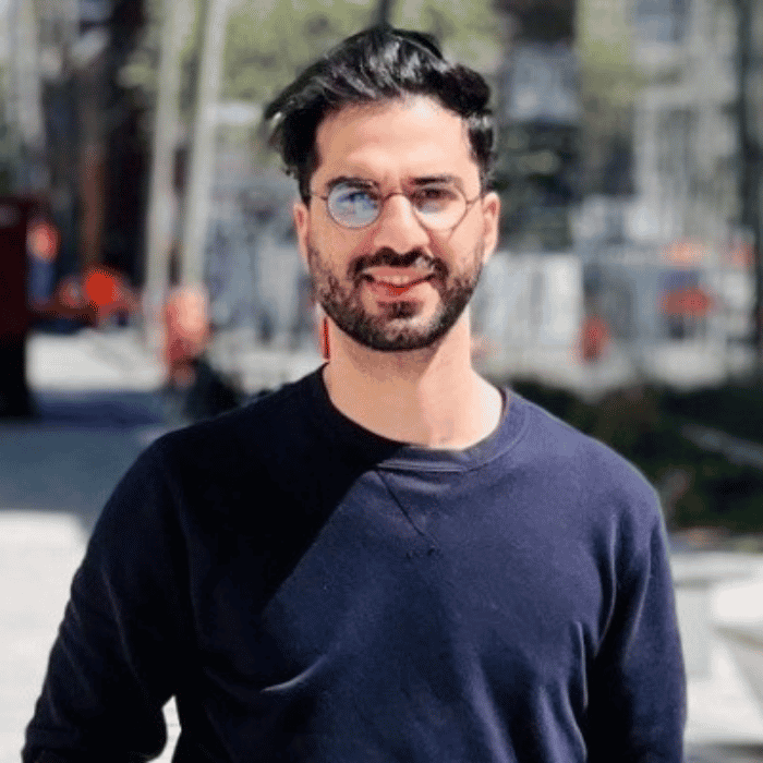 Un homme souriant se tient dans un cadre urbain, portant un pull noir et des lunettes rondes, avec un fond flou de lumières.