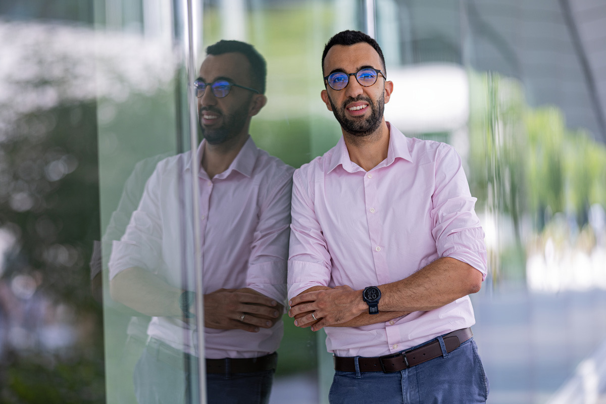 Un homme souriant en chemise rose se tient devant une vitre, reflétant à la fois sa posture assurée et un environnement moderne.