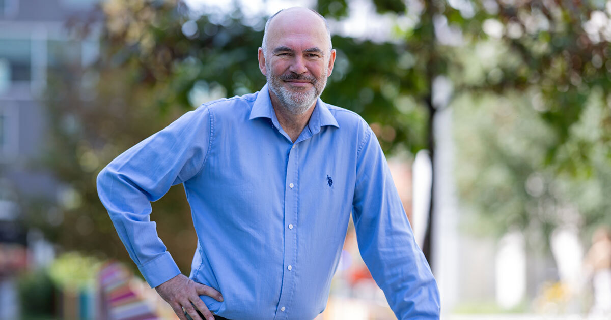 Un homme souriant, vêtu d'une chemise bleue, se tient dans un environnement verdoyant, dégageant une ambiance accueillante et professionnelle.
