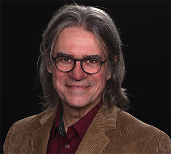 University faculty member with glasses and a warm smile in a corduroy jacket.