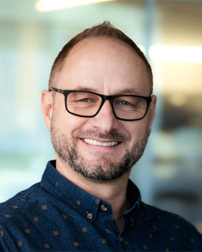 Un professeur souriant, portant des lunettes et une chemise à motifs, se tient dans un cadre professionnel moderne.