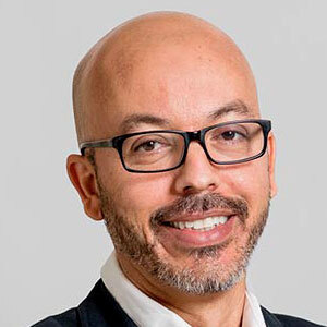 Un homme souriant, avec des lunettes, un look soigné, se tenant sur fond neutre, reflétant une ambiance professionnelle et accueillante.