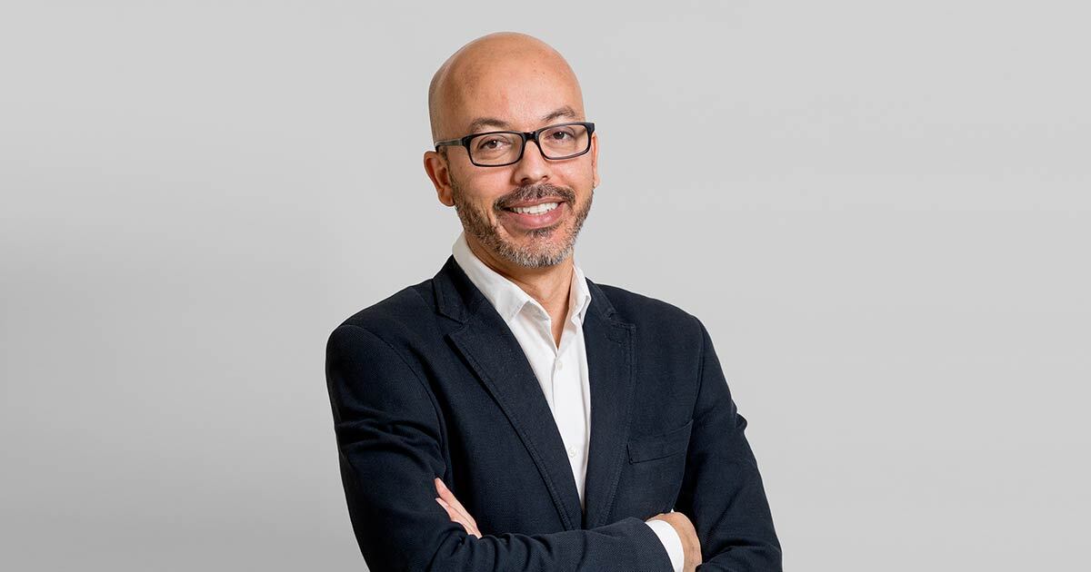 Portrait d'un homme souriant, vêtu d'un costume élégant, avec des lunettes, se tenant debout dans un environnement neutre.