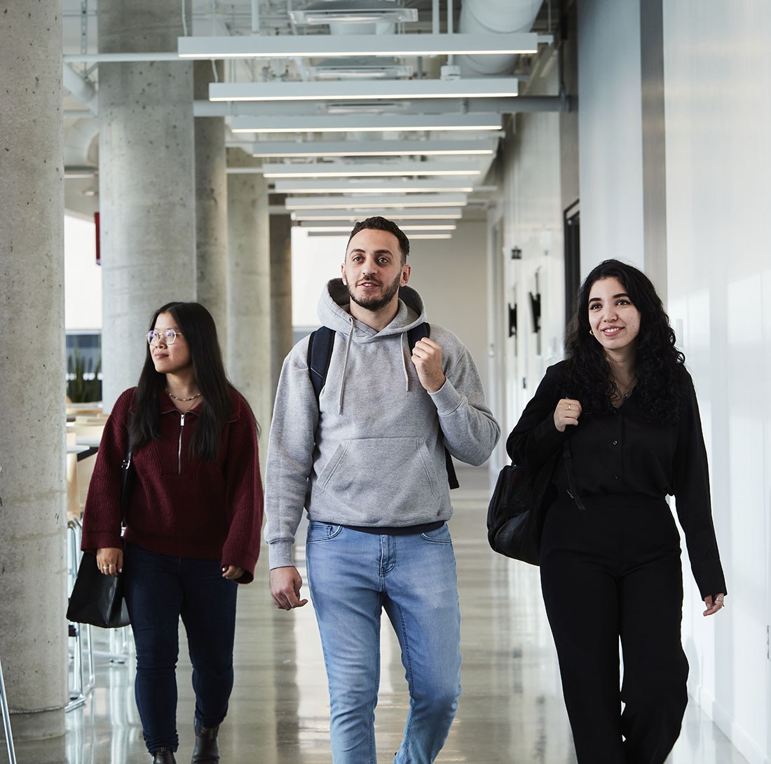 Trois étudiants marchent dans un couloir moderne, échangeant des sourires et discutant, reflétant une ambiance conviviale et dynamique.
