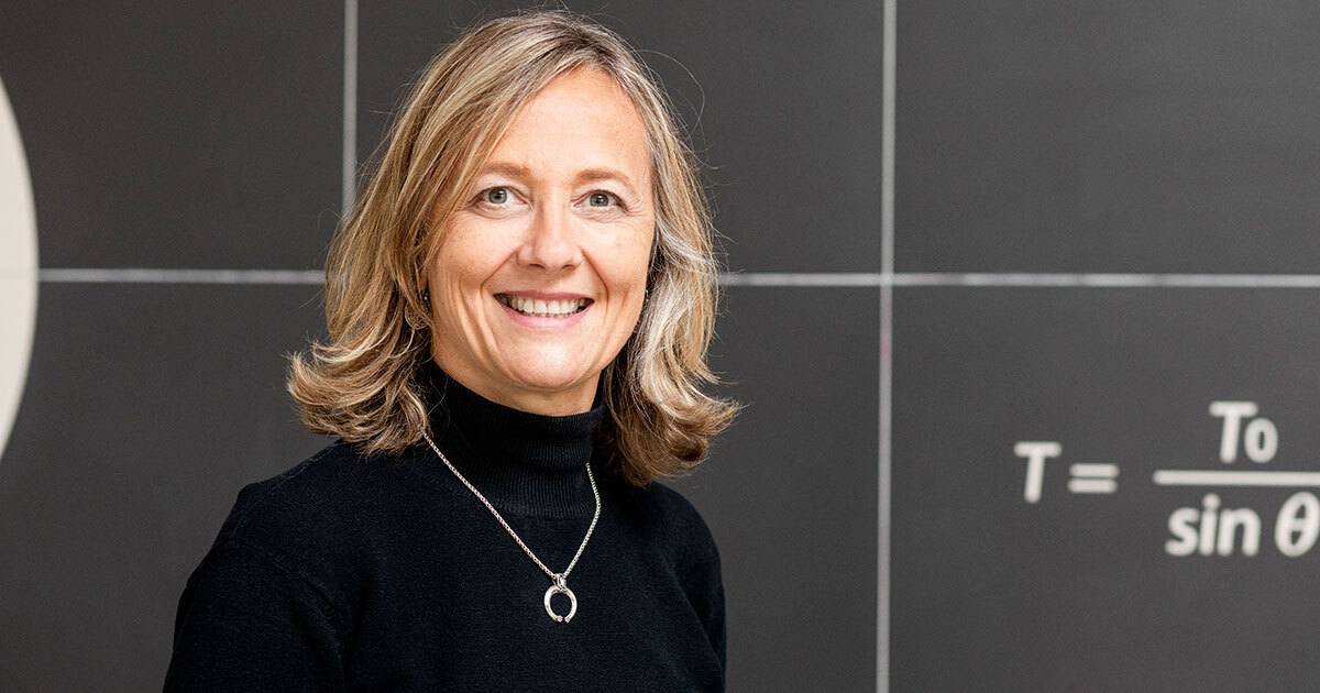 Professeure souriante, vêtue d'un haut noir, devant un mur illustrant des équations mathématiques. Ambiance académique inspirante.