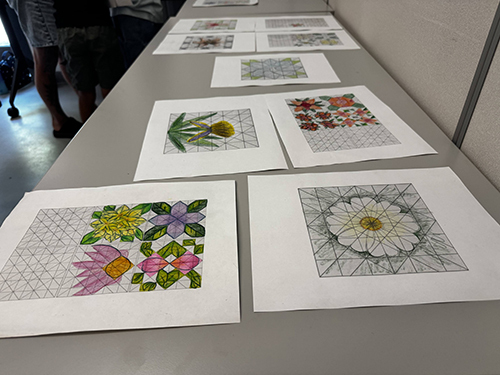Des esquisses colorées de fleurs et motifs géométriques sont disposées sur une table, illustrant le travail artistique des étudiants.