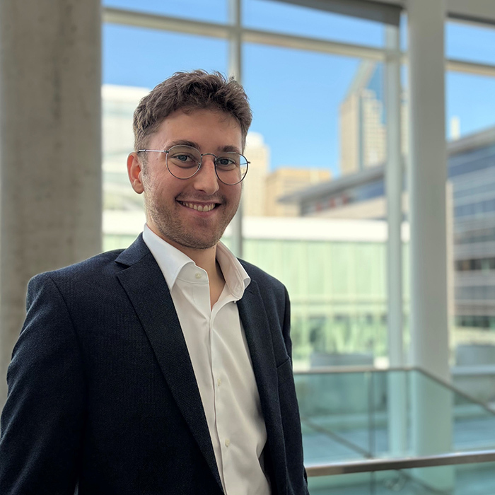 Un étudiant souriant portant un costume se tient devant une grande fenêtre, avec des bâtiments modernes en arrière-plan.