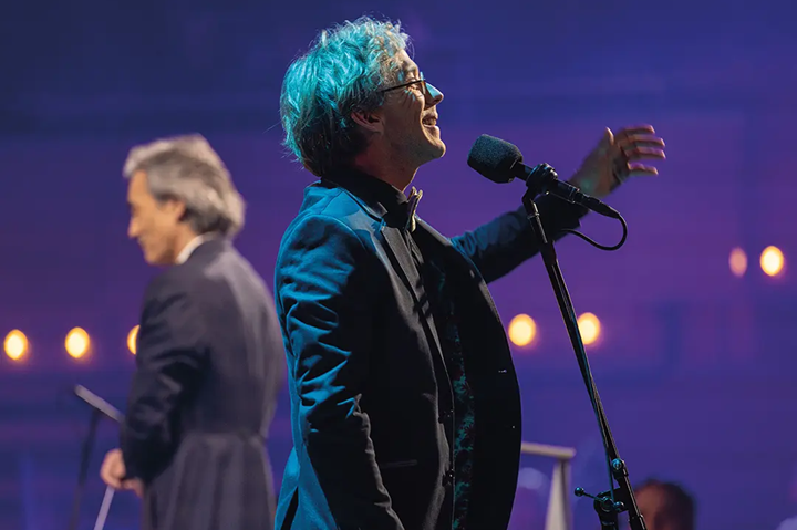Un homme en costume s'exprime avec enthousiasme devant un microphone, éclairé par des lumières tamisées, dans une ambiance conviviale.
