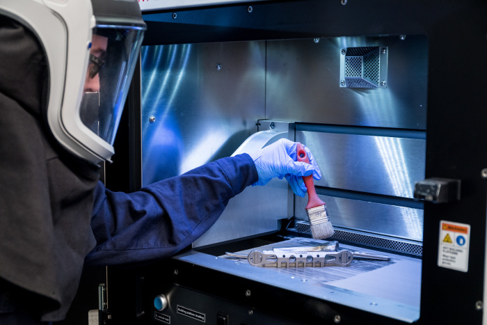 Un professionnel en tenue de protection utilise un pinceau pour manipuler des pièces dans un environnement technologique sécurisé.