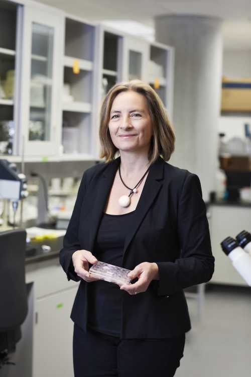 Une femme en lab coat sourit dans un laboratoire, tenant un échantillon, entourée d'équipements scientifiques modernes.