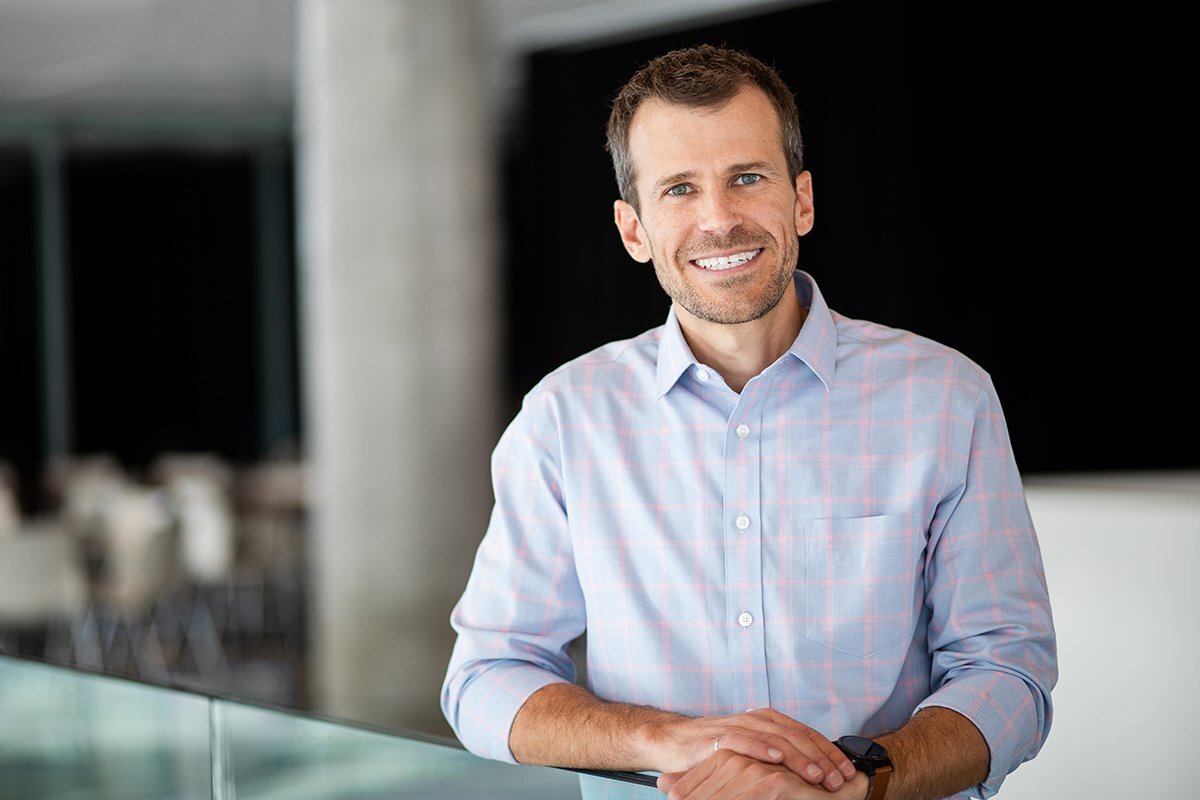 Un homme souriant se tient près d'un verre, vêtu d'une chemise à carreaux. L'ambiance est moderne et accueillante.