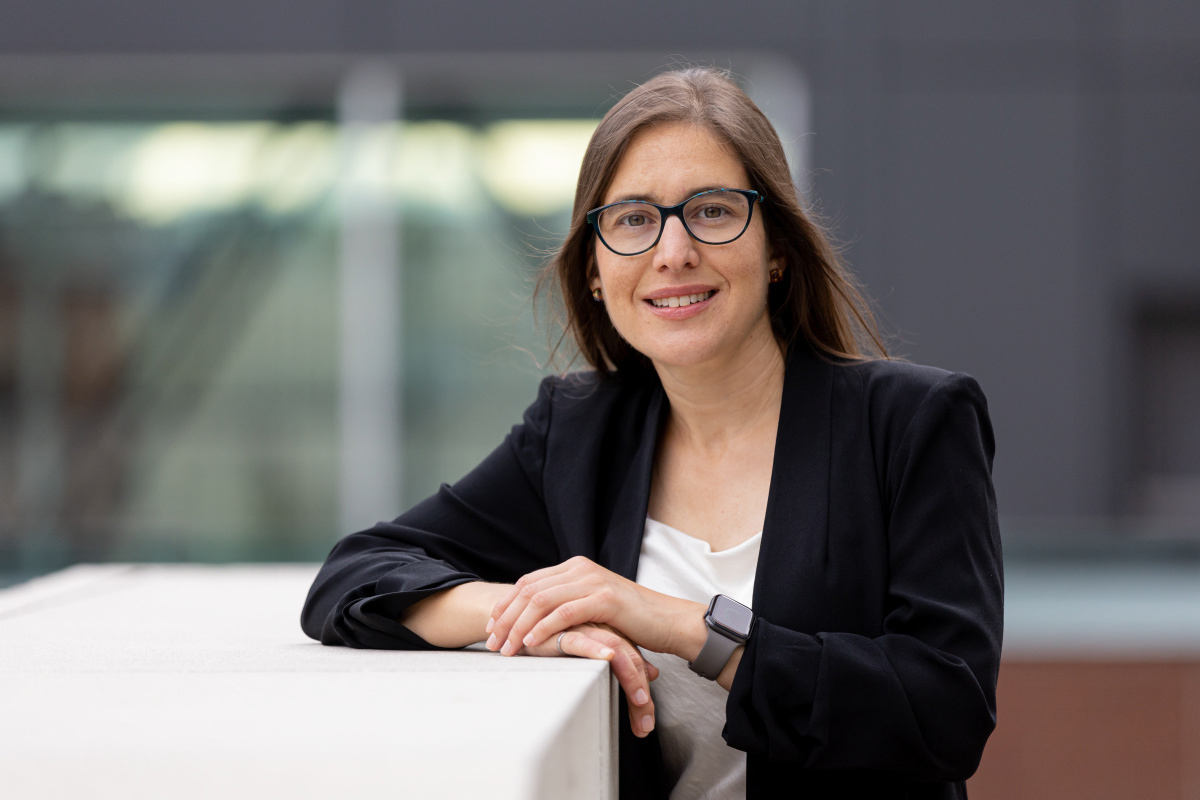 Professeure souriante avec blazer noir devant un bâtiment moderne.