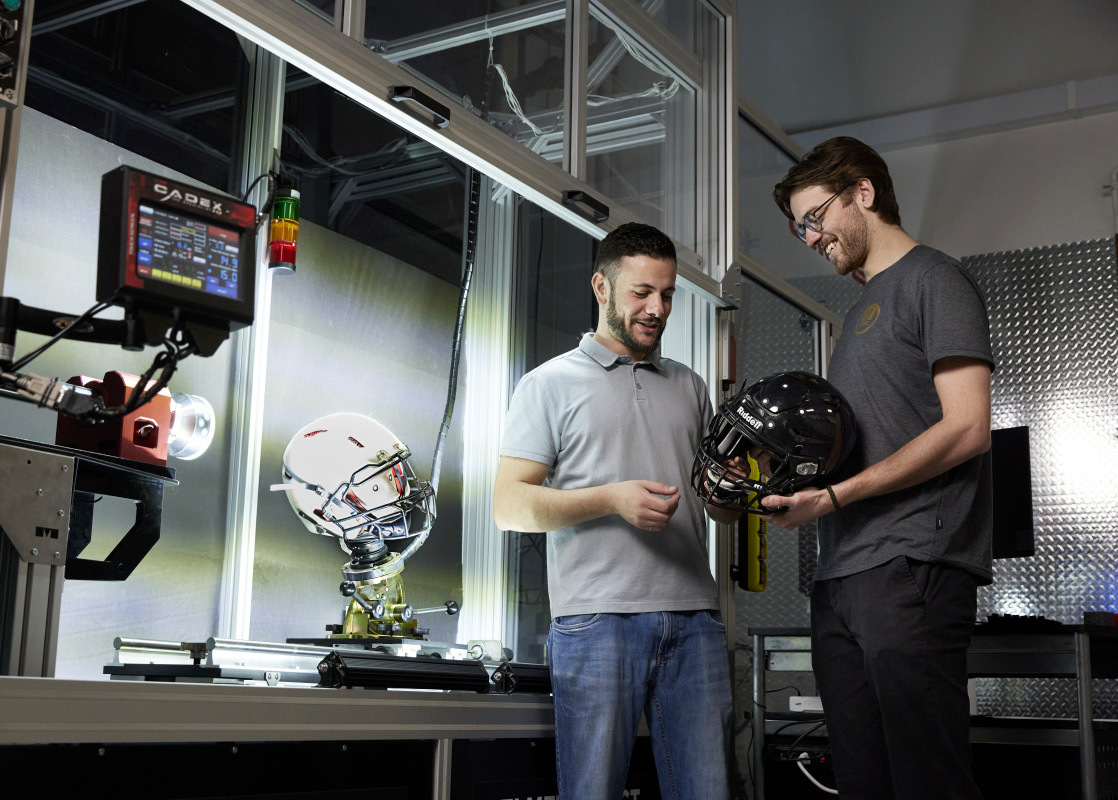 Deux chercheurs échangent autour de casques de sécurité, illustrant l'innovation en technologies de protection sportive.