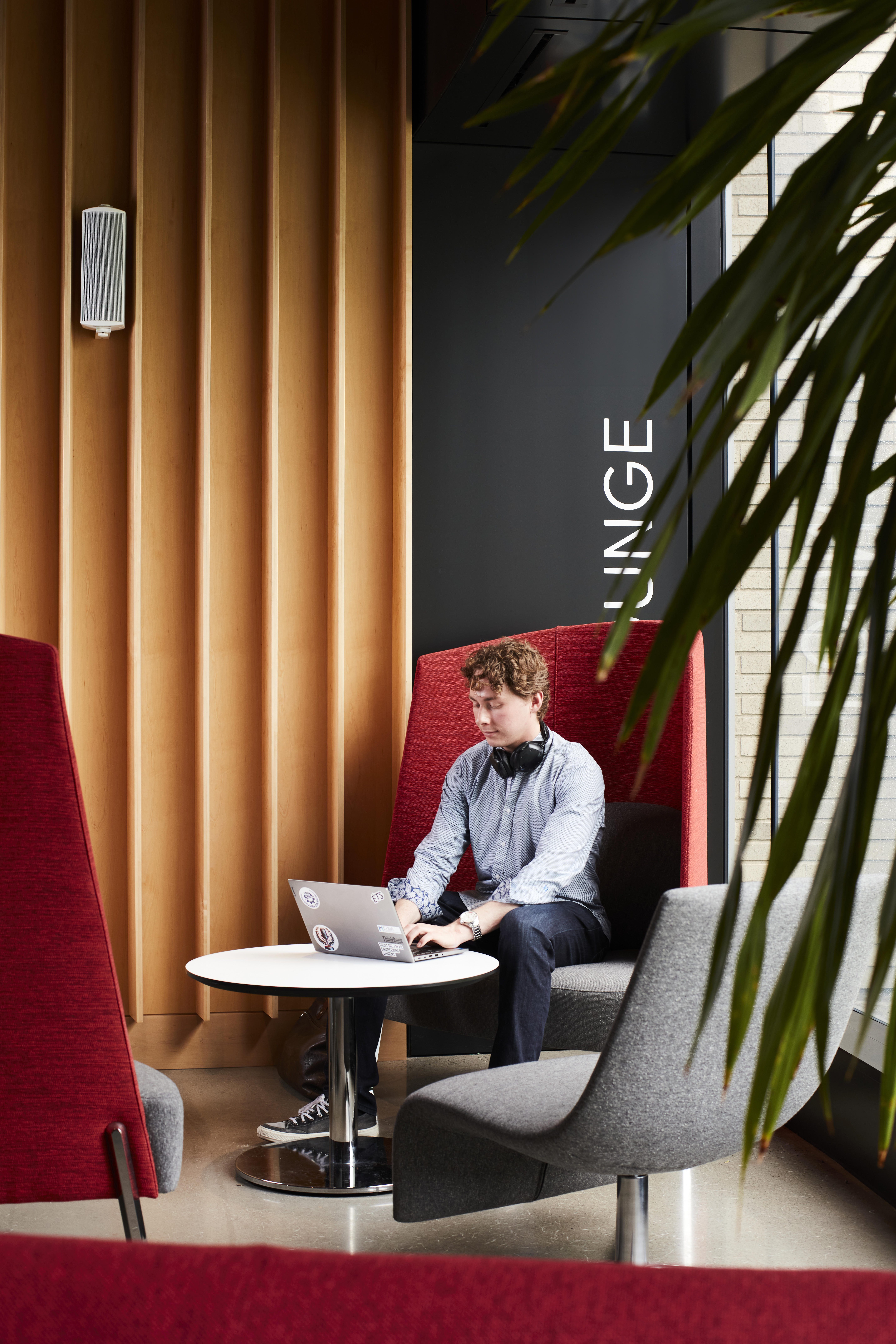 A student works intently on a laptop in a modern lounge area, surrounded by vibrant furnishings and greenery.