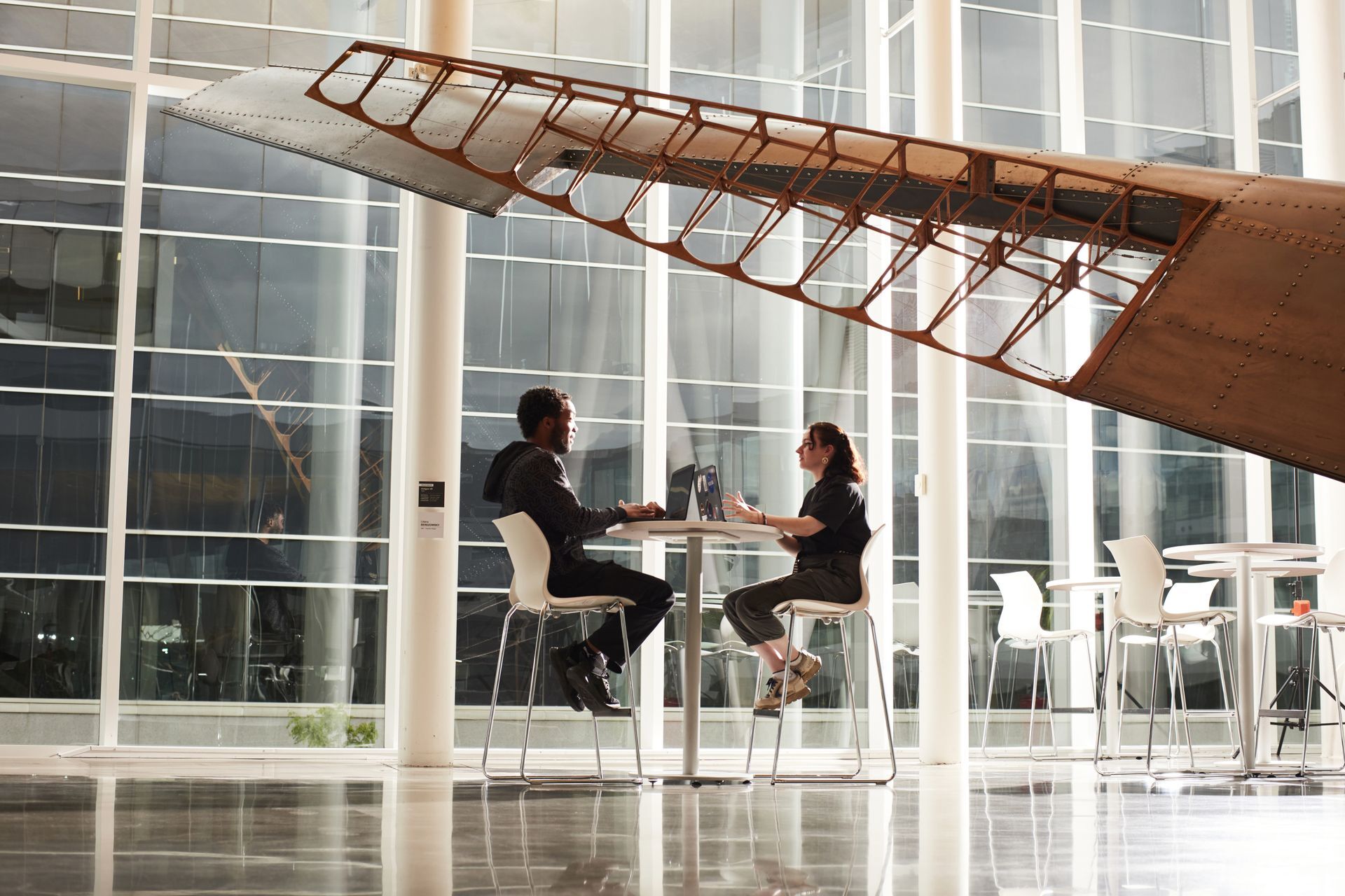 Deux étudiants discutent autour d'une table, sous une aile d'avion, dans un espace moderne et lumineux d'une institution technologique.