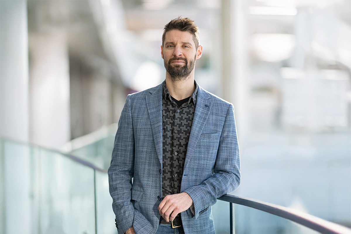 A professional individual stands confidently near a glass railing, showcasing a stylish blazer and a patterned shirt against a modern backdrop.