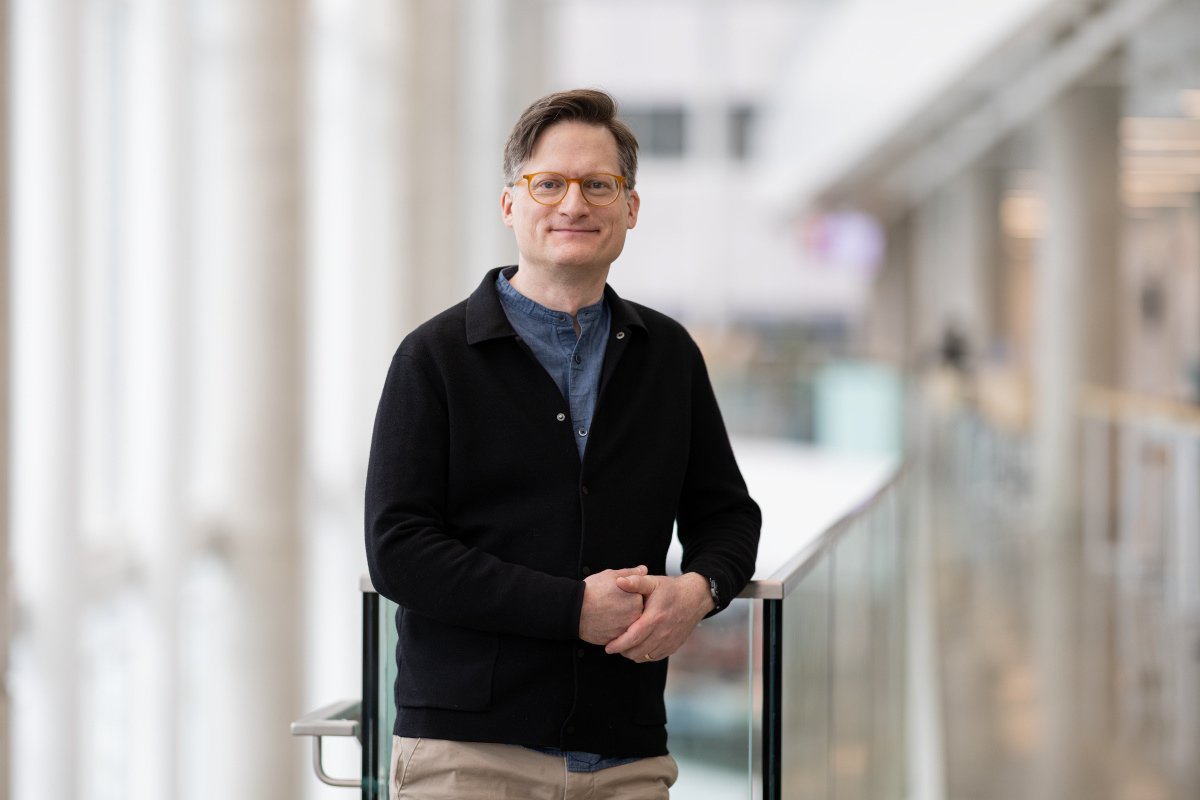 A professional smiling individual stands confidently near a glass railing in a modern, well-lit academic environment.