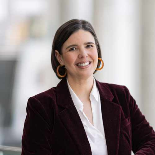 Portrait d'une professionnelle souriante, vêtue d'un blazer bordeaux, se tenant dans un cadre moderne lumineux.