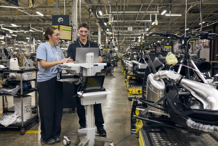 Deux étudiants échangent des idées devant un poste de travail dans une usine, entourés de machines en cours de fabrication.