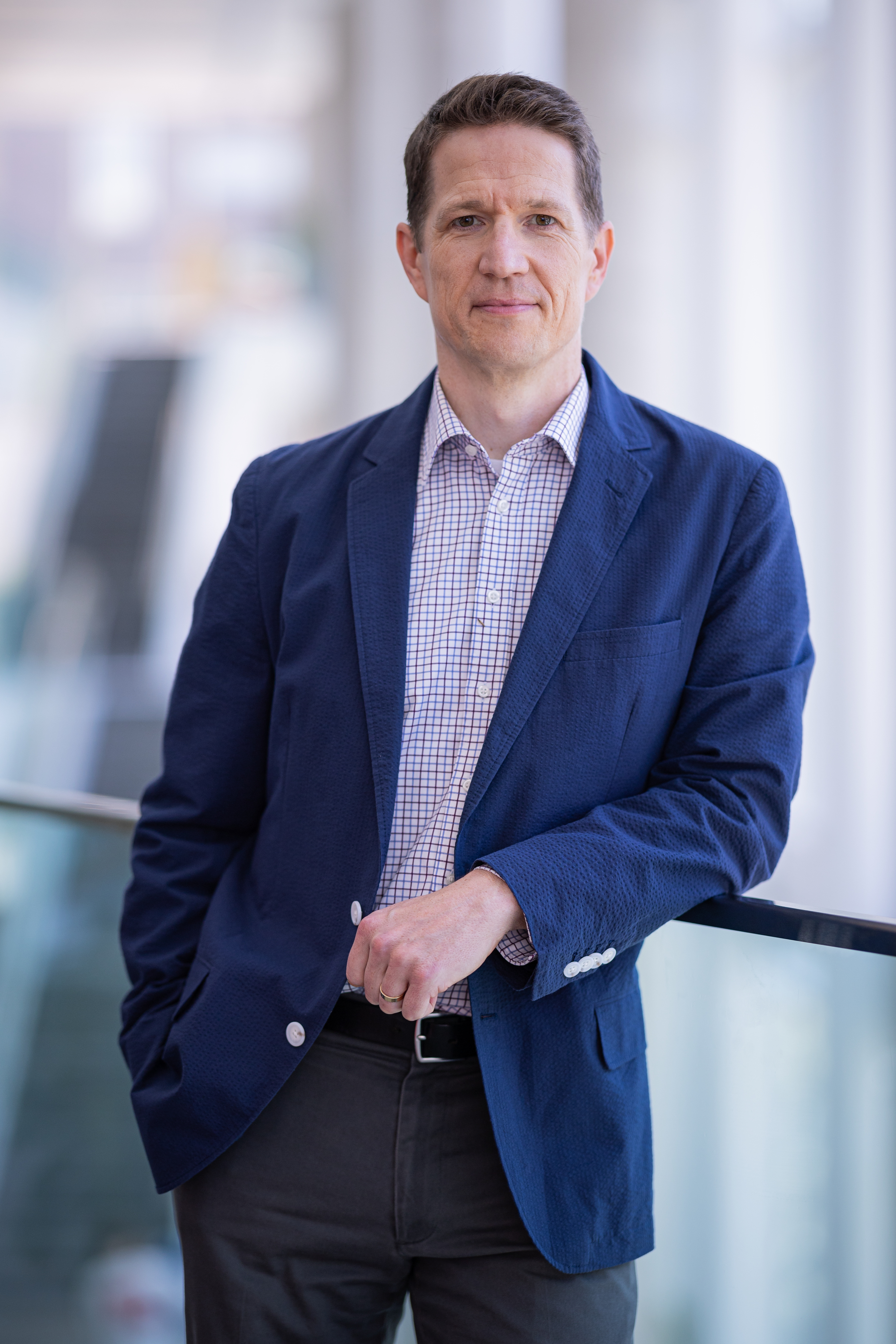 Professeur souriant, vêtu d'un blazer bleu, se tenant près d'une rambarde dans un environnement moderne et lumineux.