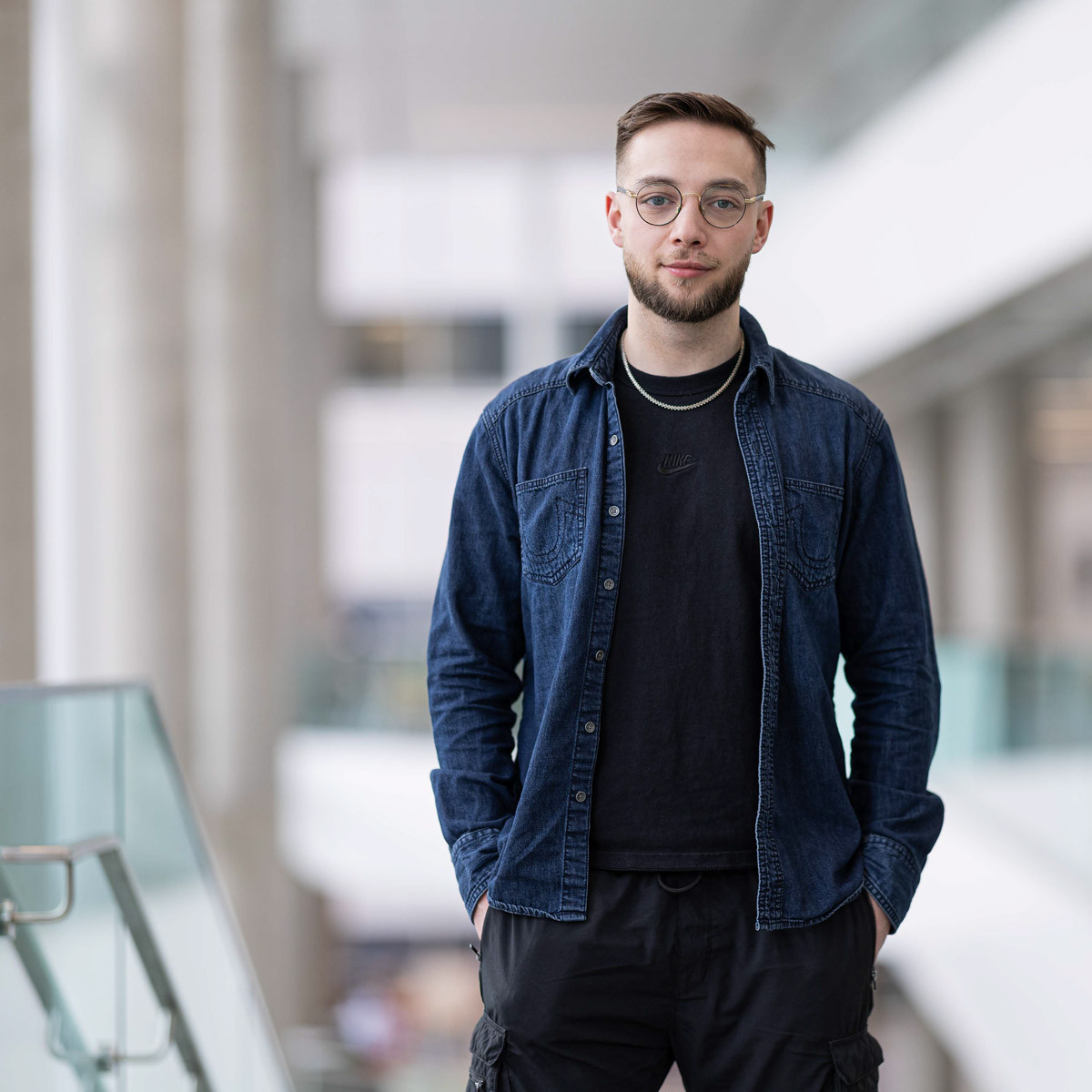 Étudiant en milieu moderne, vêtu de manière décontractée, avec un regard déterminé et une posture confiante dans un environnement universitaire.